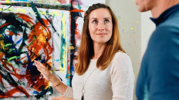 Frau deutet auf ein von ihr gemaltes Kunstwerk